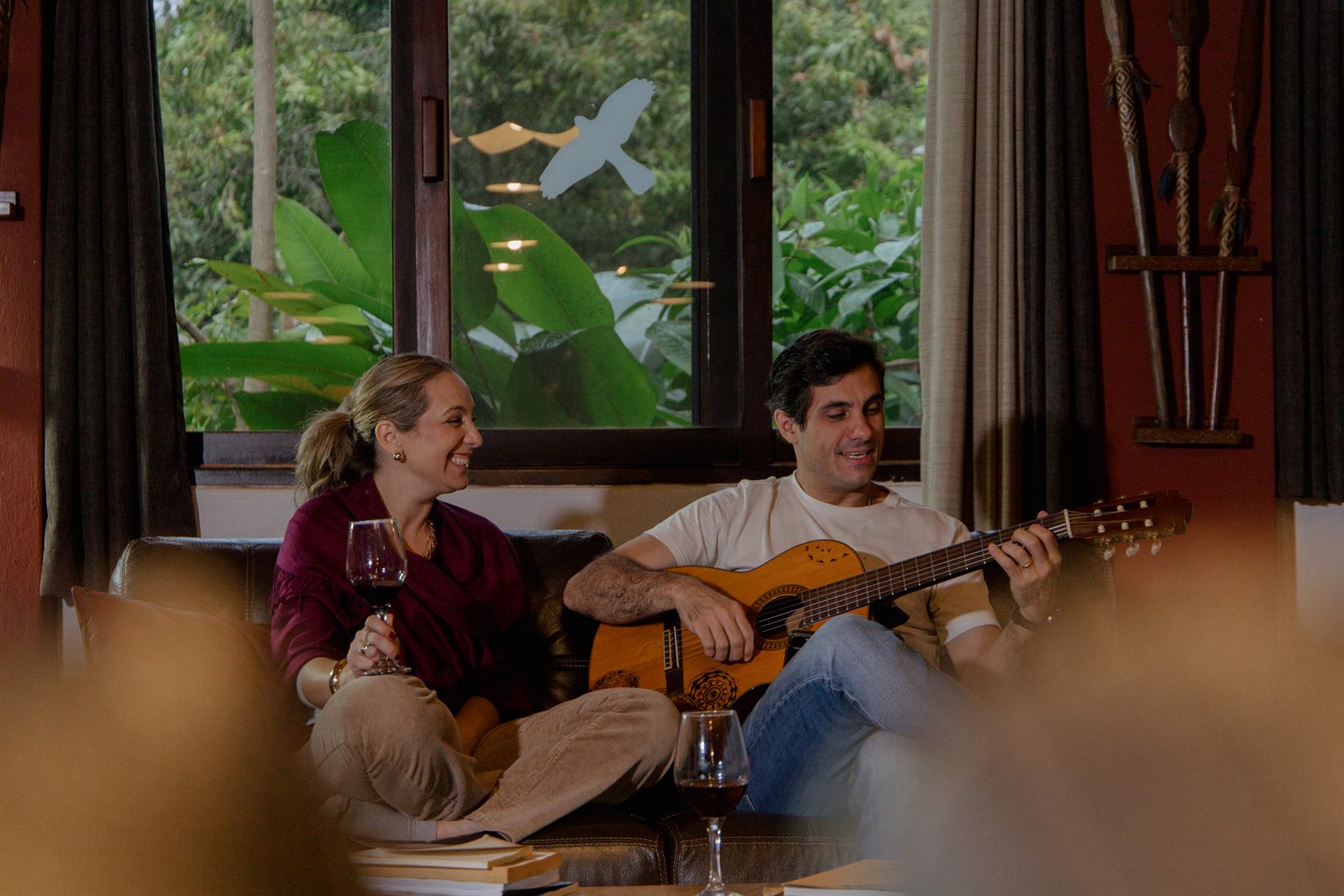 Casal no sofá com violão e taças de vinho, diante da janela para o jardim